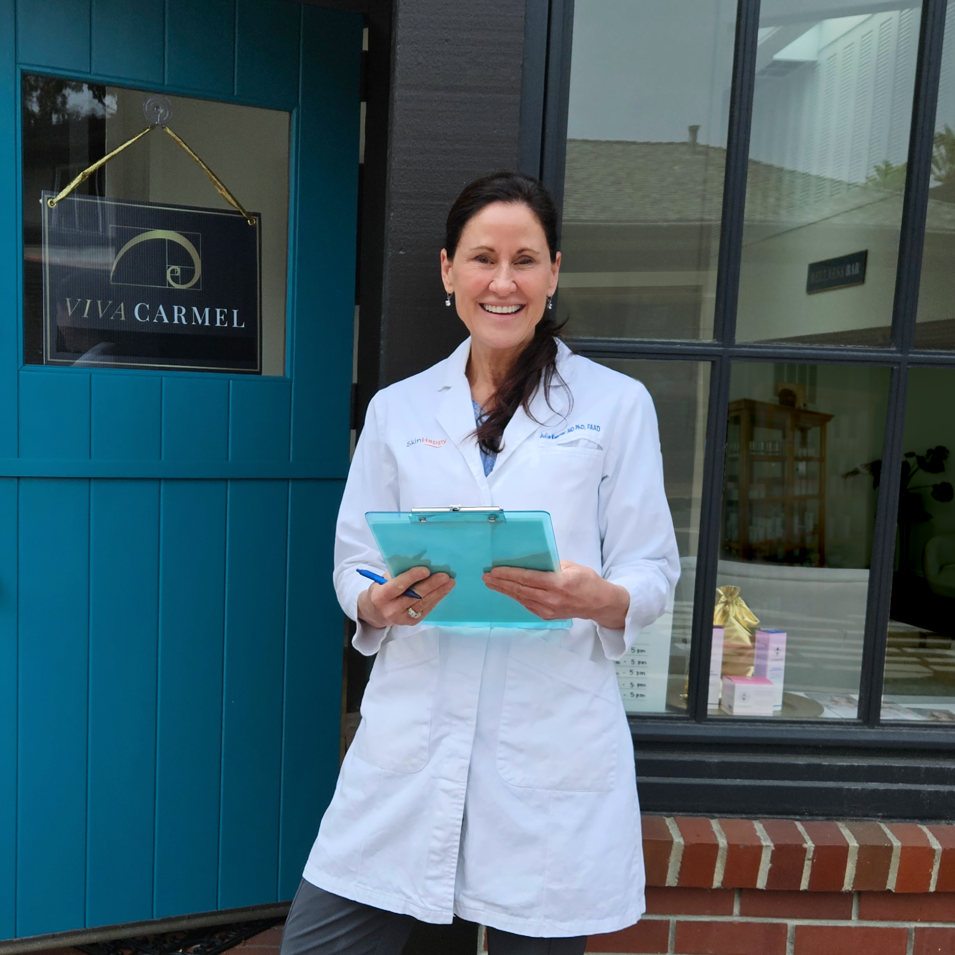 Dr. Julie Kenner, in a white coat and holding a clipboard, standing in front of a blue door with a Viva Carmel sign.