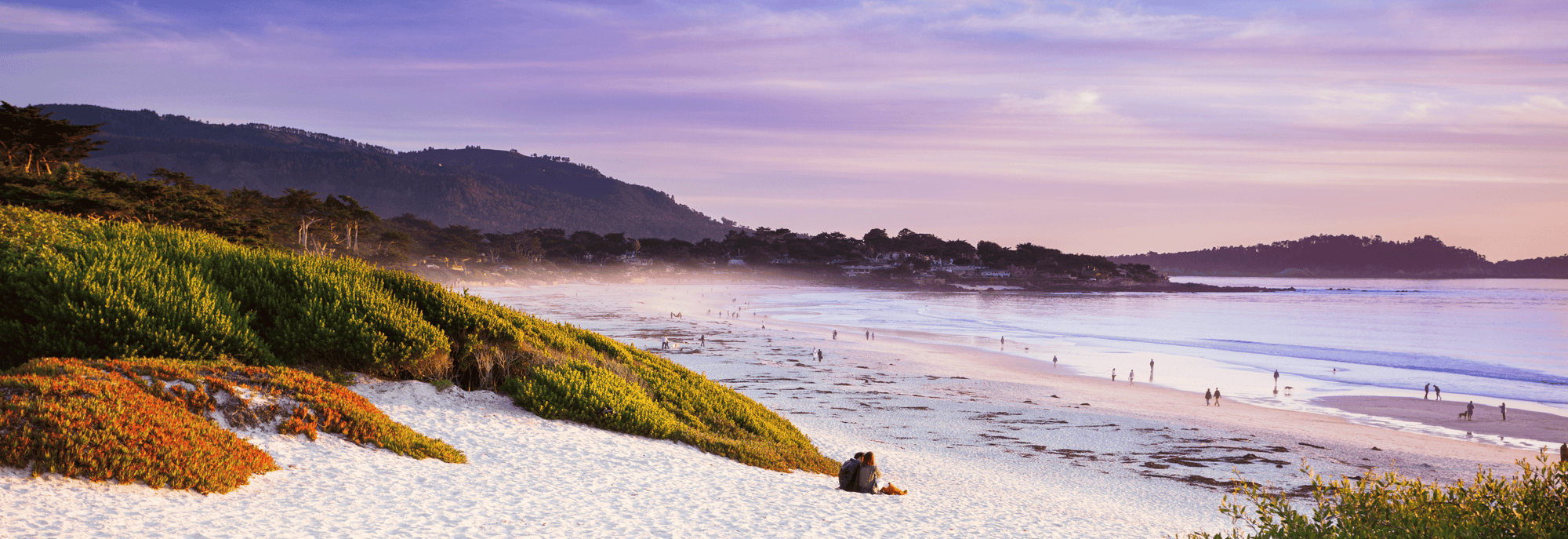A scenic coastal landscape of Carmel-by-the-Sea showing a sandy beach, ocean water, distant hills, and purple-pink sky at sunset.
