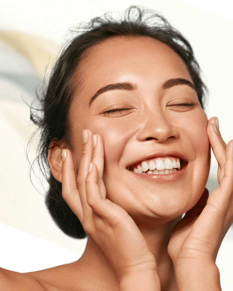 A close-up of a smiling woman with closed eyes and hands on her cheeks, showing radiant skin, against a soft light beige background.