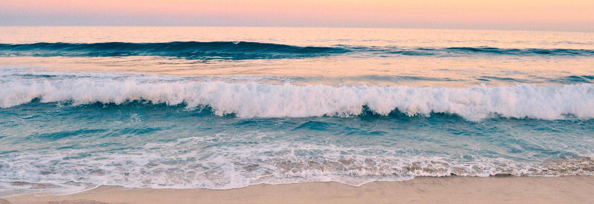 A scenic beach seascape at sunset with turquoise ocean waves rolling onto a sandy shore under a soft peach and pink sky.