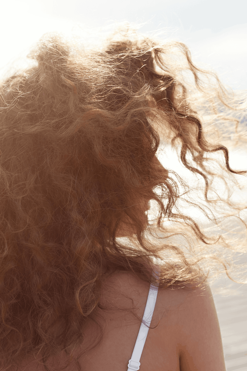 A person with curly hair blowing in the wind at the beach.
